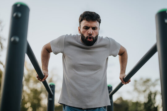 A determined man works out on parallel bars in an outdoor park setting, concentrating intently on his exercise routine. The clear sky and greenery around him create a perfect fitness environment.