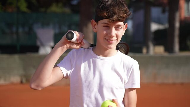 A cute, handsome teenage tennis player walking on a tennis court with his racket and smiling.