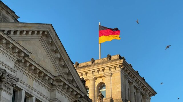 The German flag waves atop the historic reichstag building with classical architectural elements against a clear blue sky