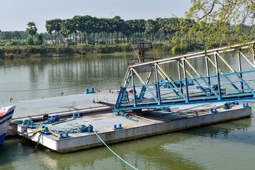 Fototapeta premium This image features a metallic blue floating pontoon bridge or ferry dock on a calm Ganges River