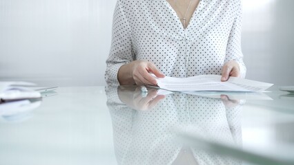 Businesswoman in white dotted blouse is going through paperwork while holding documents at a glass desk in a well lit, contemporary office setting. Business people concept