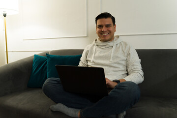 Smiling male freelancer relaxing on home sofa, typing on laptop during remote work session