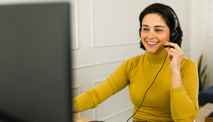 Cheerful sales representative wearing headset talking with customer and working on computer from...