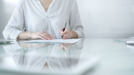 Businesswoman in white dotted blouse is signing a contract at a sleek glass desk in a modern...