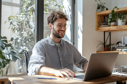 Smiling man working on laptop at bright, sunny desk.