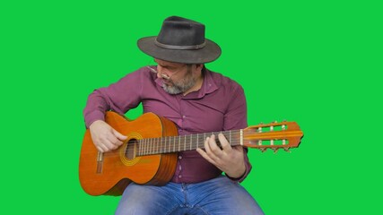 A man in a hat sits and plays an acoustic guitar against a green screen background. This shot is ideal for music videos, commercials, or any project needing a musician with a chroma key backdrop