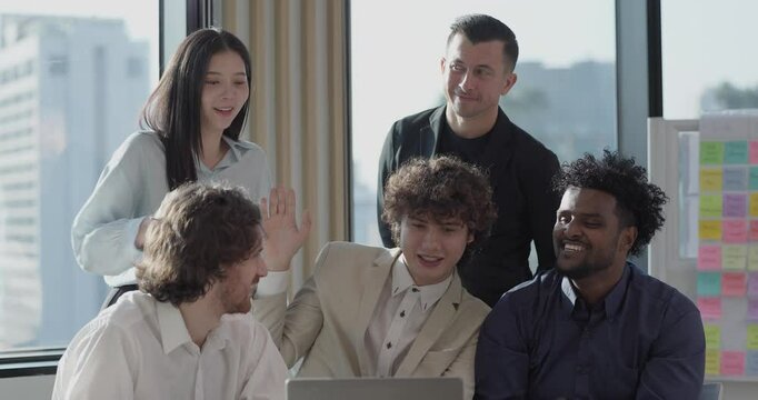 Diverse startup team engages in video conference, smiling at laptop screen. The bright workspace features a whiteboard with colorful sticky notes, reflecting teamwork, innovation, and collaboration
