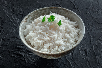 White rice, cooked. A bowl of boiled long grain rice, a simple side dish, with fresh parsley leaves, on a black slate background