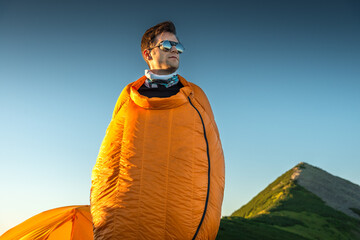 Directly below, a young man standing in a sleeping bag near a tent at sunrise, wearing sunglasses and enjoying the morning light in the mountains.