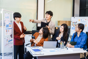 Five young Asian professionals collaborate at startup business meeting, working at desk with laptops and financial charts. They discuss strategy, funding, innovation, teamwork and problem solving.