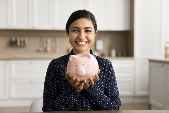 I see my future wealthy. Portrait happy smiling young woman sit at kitchen desk look at camera show you funny piggybank. Satisfied bank client keep finances on deposit use cashback program save money