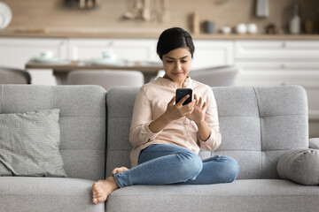 Fototapeta premium Leisure time online. Happy smiling millennial woman rest on comfy sofa at home using web app on mobile telephone text message communicate in chat enjoy watching video at social media read news in blog