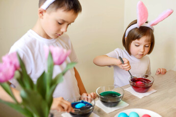 Two children, boy and toddler girl, wearing bunny ears, dyeing Easter eggs, stirring paint in glass...