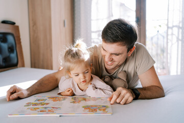 Smiling dad with a little girl lie on their stomachs on the bed and look at a book with bright...