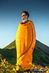 A young man with tousled hair stands wrapped in an orange sleeping bag on a rocky ridge at sunrise,...