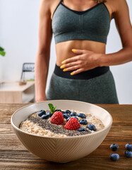 Femme sportive d&rsquo;environ 30 ans en tenue de fitness touchant son ventre, devant un bol de porridge aux flocons d&rsquo;avoine, fruits rouges et graines de chia