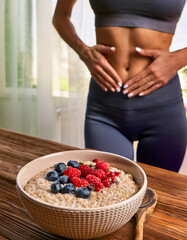 Femme sportive d&rsquo;environ 30 ans en tenue de fitness touchant son ventre, devant un bol de porridge aux flocons d&rsquo;avoine, fruits rouges et myrtilles