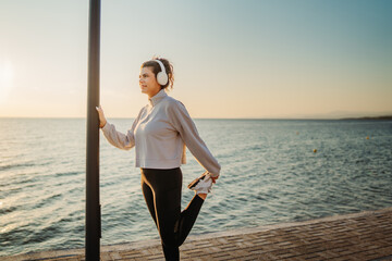 Young caucasian woman put on headphones and stretch before run on beach at sunset