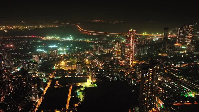 An aerial view of Mumbai's high-rise buildings and beautiful nightlights. The sky is cloudy, and there is medium traffic on the road. The cityscape is filled with modern illuminated buildings.
