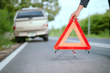 Man on roadside holds warning triangle after car breakdown during daylight near a lush green landscape