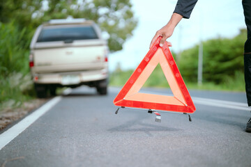 Man on roadside holds warning triangle after car breakdown during daylight near a lush green landscape