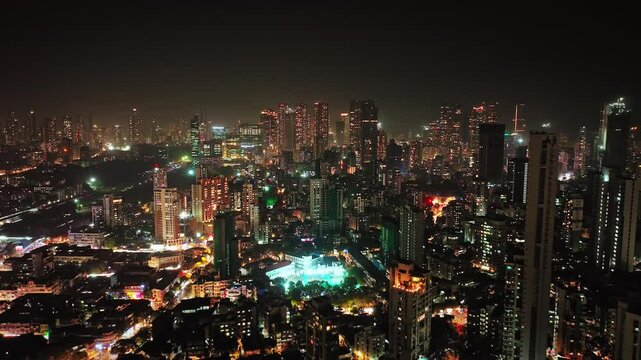 An aerial view of Mumbai's high-rise buildings and beautiful nightlights. The sky is cloudy, and there is medium traffic on the road. The cityscape is filled with modern illuminated buildings.