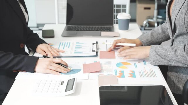 Two female business professionals in suits sit at desk, engaged in strategic discussion. They analyze financial planning, risk management, corporate growth while reviewing reports and market trends.