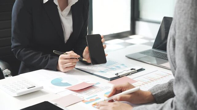 Two female business professionals in suits sit at desk, engaged in strategic discussion. They analyze financial planning, risk management, corporate growth while reviewing reports and market trends.