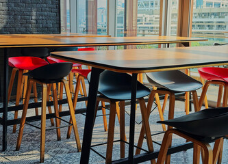 Long wooden food tables and chairs of a food court with background of long glass doors and city view, warm light in the morning, no people.