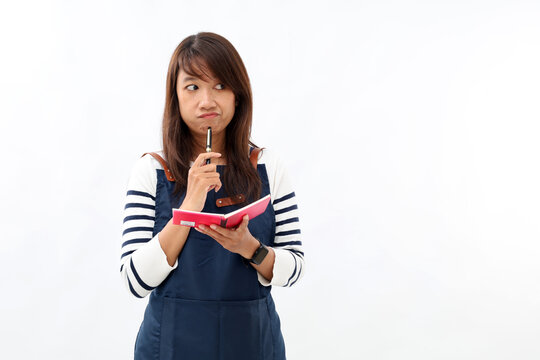 Asian barista woman in blue apron taking order, writing on menu book list, thinking with pen on chin. Isolated image on white background