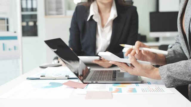 Two female business professionals in suits sit at desk, engaged in strategic discussion. They analyze financial planning, risk management, corporate growth while reviewing reports and market trends.