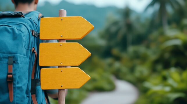 Naklejki Person with blue backpack stands before a yellow wooden signpost in a lush green tropical forest. A winding path is visible in the background