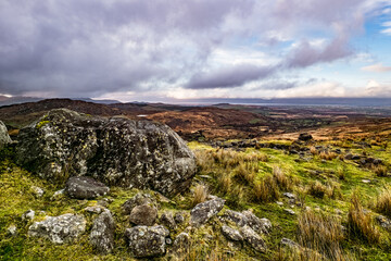 A large rock sits in the foreground, surrounded by grass and scattered stones. In the distance, rolling hills stretch out under a clear sky, showing the beauty of nature.