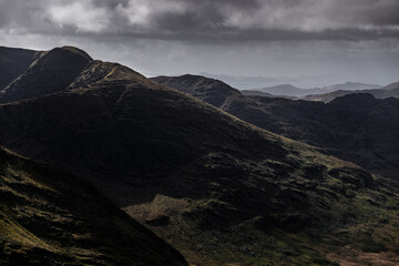The photo depicts a series of mountain ranges. The closest range shows rocky, dark brown and black...