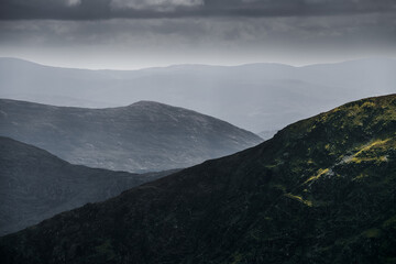 Under a soft cloudy sky, lush green mountains in varying shades create a captivating view. The landscape is peaceful and tranquil. It invites exploration.