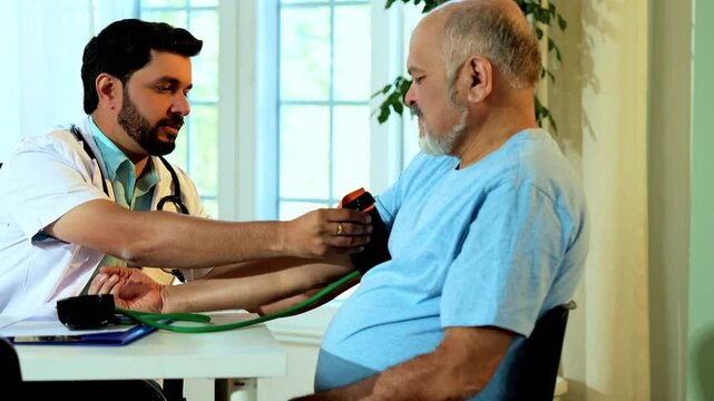 Indian Asian male doctor checks blood pressure of senior patient in OPD using a sphygmomanometer, ensuring accurate diagnosis and quality healthcare in a cozy clinic with reassuring results.