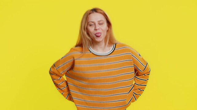 Caucasian blonde woman in blouse showing tongue making faces at camera, fooling around, joking, aping with silly face, teasing, bullying, abuse. Young adult girl isolated on yellow background indoors