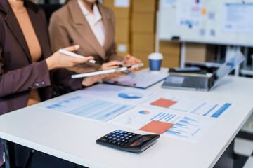 Two female business professionals in suits sit at desk, engaged in strategic discussion. They...