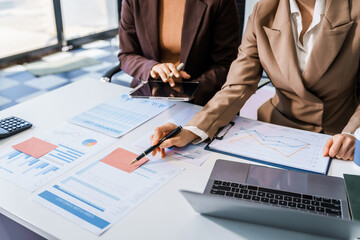 Two female business professionals in suits sit at desk, engaged in strategic discussion. They...