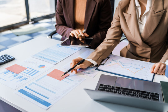 Two female business professionals in suits sit at desk, engaged in strategic discussion. They analyze financial planning, risk management, corporate growth while reviewing reports and market trends.
