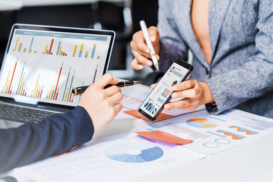 Two female business professionals in suits sit at desk, engaged in strategic discussion. They analyze financial planning, risk management, corporate growth while reviewing reports and market trends.