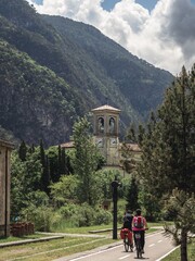 Two cyclists ride along the Alpe Adria biking route, passing a historic church tower surrounded by lush greenery and towering mountains in Italy. A peaceful and scenic travel destination.