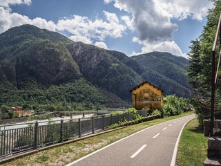An old yellow train station in Chiusaforte, Italy, alongside the Alpe Adria biking route. Green mountains and a river create a stunning natural backdrop under a partly cloudy sky.