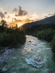 A scenic view of a fast-flowing mountain river surrounded by lush greenery and rugged hills at sunset in northern Italy. The golden light reflects on the water, creating a tranquil yet dynamic scene.