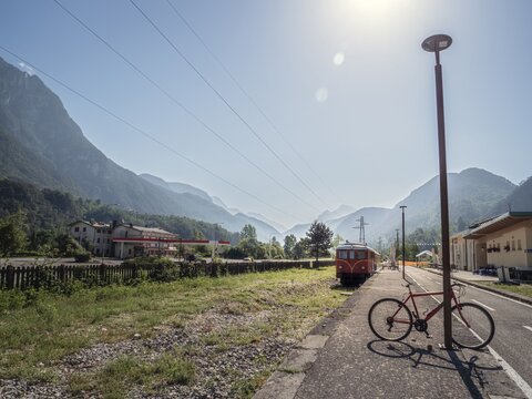 A red bicycle leans against a lamppost at a rural train station with a vintage red train in the background. The scenic mountain landscape and power lines add depth to the peaceful setting.