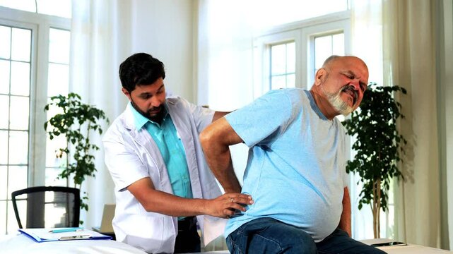 Indian Asian male physiotherapist examining senior retired patient with chronic back pain in a lavish clinic, ensuring precise diagnosis and effective rehabilitation for pain relief and improved mobil
