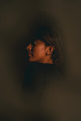 A woman with dark hair and brown eyes is standing in front of a wall. The image has a moody and...