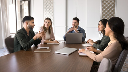 Five multiethnic professionals gathered around conference table listening speech of young skilled Arabian male colleague, make speech, presents idea or point during group meeting, briefing with staff