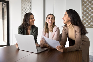 Three attractive multiracial women colleagues gathered in office with laptop, laughing, share new...