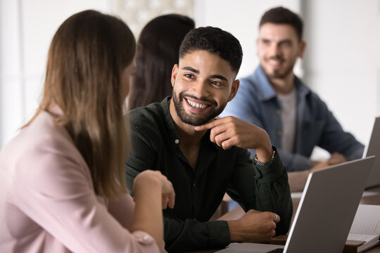 Young smiling Arabian man listening woman colleague seated together at shared table with laptop in coworking space, participants involved in joint task discussion, teamwork and collaboration in office